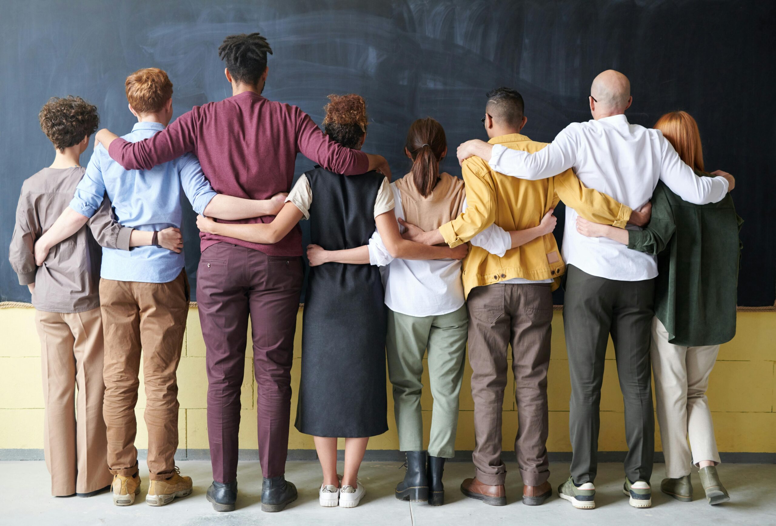 Group of people embracing with their backs to the camera. They are standing facing a blank blackboard.